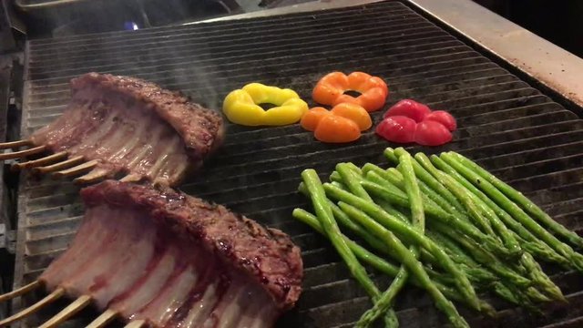 Close up of lamb and vegetables on grill with chef flipping meat with tongs.
