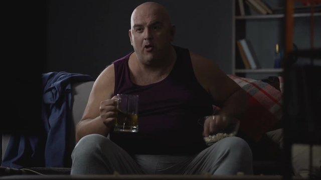 Plump guy watching TV with popcorn and beer, unemployment and laziness problems