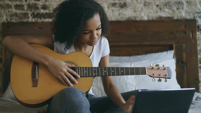 Young curly mixed race girl concentraing learning to play guitar using laptop computer sitting on bed at home