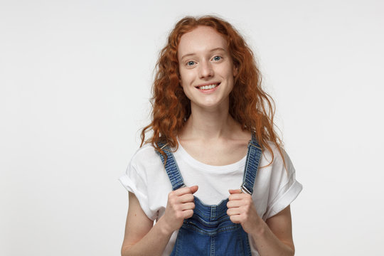 Indoor portrait of young beautiful redhead European female isolated on white background holding straps of jumpsuit in hands, showing inspired smile to her mate with relaxed and friendly look