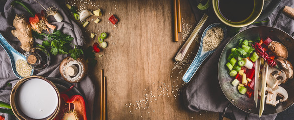 Vegetarian Asian cuisine ingredients for stir fry with chopped vegetables, coco milk, spices,chopsticks and wok pot on rustic wooden background, top view, banner. Chinese or Thai food cooking