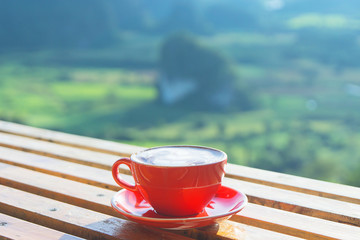 The cappuccino coffee in red cup put on the wooden table with beautiful mountain landscpe scenery and sun light in the morning.
