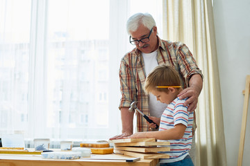 Portrait of nice senior man helping little boy hammering nails while making wooden model together standing against window at home, copy space