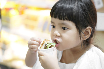 Asian children cute or kid girl taste and eating sandwich or hamburger for delicious or not delicious food in the supermarket at department store