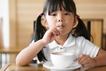Asian children cute or kid girl with delicious hot cocoa or chocolate drink in white cup are full and spoon at mouth and sloppy for breakfast in morning on table at home or cafe restaurant and looking