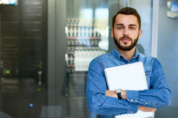 Portrait of beraded systems administrator posing holding laptop and looking at camera standing against server cabinet, copy space - obrazy, fototapety, plakaty