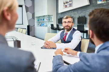 Portrait of barded mature businessman listening  to partners sitting at meeting table in board room while negotiating deal