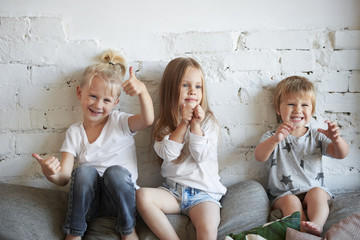 Candid shot of three siblings posing in living room, sitting on top of sofa at white brick wall:...