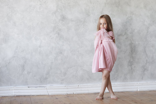 Full length portrait of beautiful barefooted timid European little girl with long loose hair standing on wooden floor at home, crossing her legs, feeling shy while posing in new pink fancy dress