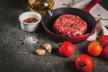 Raw organic beef meat burgers cutlets  with spices, thyme, tomatoes, olive oil in frying pan on black background, copy space