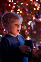 Little toddler boy holding candle with colorful lights from Christmas tree on background.