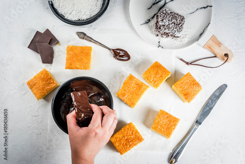 Australian cuisine. Preparation of traditional Australian dessert Lamington: biscuit in chocolate with coconut shaving powder. Top view, female hands in the frame. 