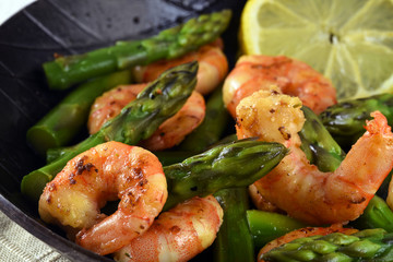 fried prawns or shrimps with  green asparagus peaks and a lemon slice in a black iron pan, close up