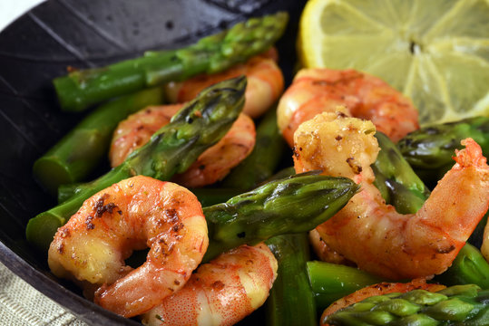 fried prawns or shrimps with  green asparagus peaks and a lemon slice in a black iron pan, close up