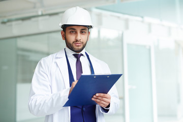 Young worker in whitecoat and helmet making notes in document