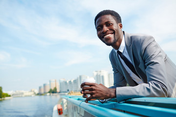 Smiling businessman with glass of coffee looking at camera by seaside