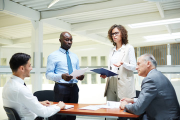 Mature and young businessmen discussing reports of their intercultural colleagues at briefing