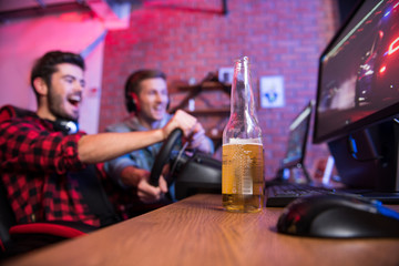 Playful mood. Low angle close-up focus bottle of light beer is on computer table. Joyful young gamers with headphones are playing car racing video game using steering wheel in background