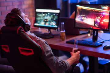 Feeling tired. Back view of relaxed young gamer in headphones is resting in chair and drinking beer...