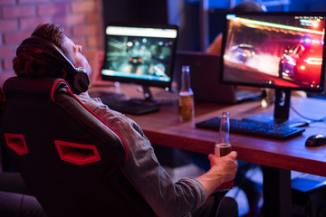 Feeling tired. Back view of relaxed young gamer in headphones is resting in chair and drinking beer while enjoying car racing video game. Selective focus © Yakobchuk Olena