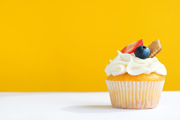 Cupcake with whipped cream, fresh strawberry, blueberry and cookie on yellow white background. The image with copy space. Background for the confectionery menu, cards, greetings, birthday invitations.