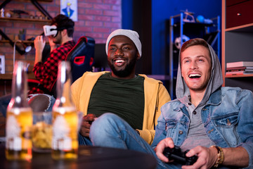 Obraz na płótnie Canvas Adult toys. Joyful best international friends are using video game console and sitting on sofa while expressing excitement. Cute guy is wearing VR glasses in background. Bottles of beer in foreground