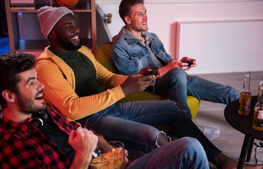 Side view of cheerful young stylish men are relaxing on sofa while using home video game console....