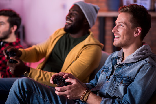 Adult party. Joyful fashionable attractive guys are enjoying play station. They are sitting on sofa and expressing gladness while holding joystick and looking at monitor. Selective focus