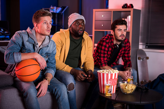 So exciting. Three young interested guys are sitting on sofa and expressing exhilaration while watching sport game with opened mouthes. Friends are eating popcorn and drinking beer - Powered by Adobe