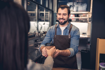 Portrait of smiling bearded worker holding out hand with mug of beverage to woman in modern cafe....