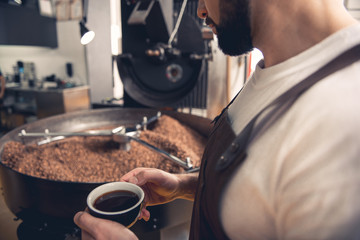 Serious bearded man tasting mug of coffee while looking at preparing beans in special roaster machine. Relax concept. Close up