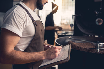 Serene bearded man writing information about roasting beans in cooler cylinder of special machine....