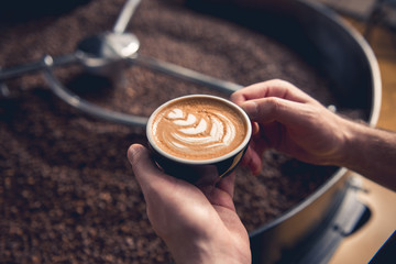 Focus on close up male hands holding cup with delicious cappuccino. Coffee grains situating in cooler machine at factory. Break concept