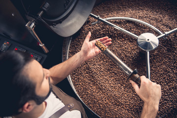 Top view serious bearded worker near coffee roaster controlling level of grain roasting. Revise...