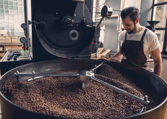 Side view smiling unshaven worker watching at coffee beans situating in grain chiller. Industry concept