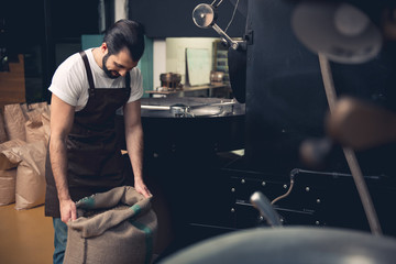 Side view cheerful bearded master holding hard bag with coffee beans while standing near technical equipment in room. Job concept