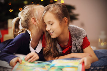 Two cute little sisters reading story book together under Christmas tree