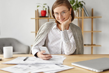 Delightful cheerful woman uses smart phone for entertainment and work, sits at office table with many documents, going to have coffee break. People, business, communication and career concept