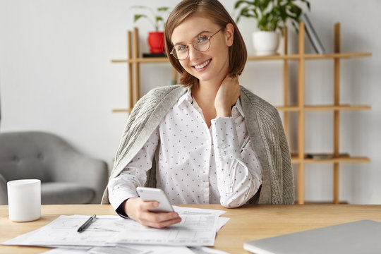 Smiling female accounter works at financial data, surrounded with many sheet of papers and modern electronic gadgets, happy to recieve message from boyfriend, invites her have supper in cafe