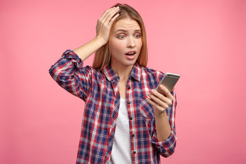 Shocked woman with long hair, keeps hands on head, looks into screen of mobile phone, recieves message remainding her about necessary thing to do, forgot about it completely, isolated on pink wall