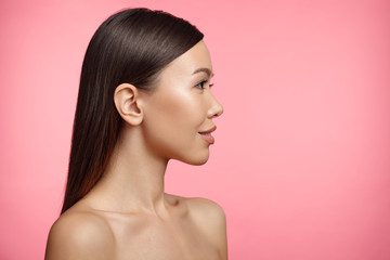 Sideways portrait of naked brunette young female with healthy skin, looks aside, isolated on pink background. Woman wears no clothes. Beauty, health concept. Adorable girl poses in studio. Feminity