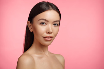 Beautiful young woman with Asian appearance, poses naked, has healthy clear skin, stands over pink background. Head and shoulders of extraordinary female. Skin care concept. Perfect naked body