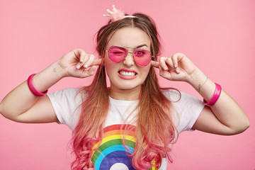 Irritated pinup girl plugs ears as hears tiresome loud noise, can`t stand hushing sound, wears t shirt with rainbow print, has pink hair tips, poses in studio. Emtional chick female bothered with buzz