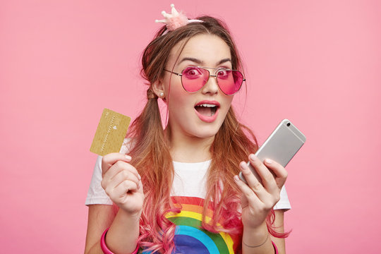 Beutiful woman holds smart phone and credit card, uses for e commerce or e payment, isolated over pink background. Attractive young female model purchases online. Pinup girl holds mobile banking