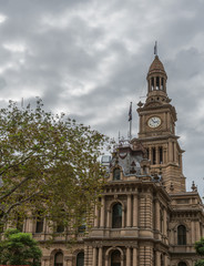 Sydney, Australia - March 25, 2017: View on corner of brown stone historic monumental City Hall with clock tower under heavy sky and tree in foreground.