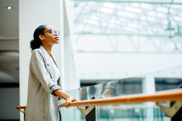 young african business woman in glasses dressed in gray suit holding on handrail when looking up and thinking indoor at modern business center. People lifestyle concept. - Powered by Adobe