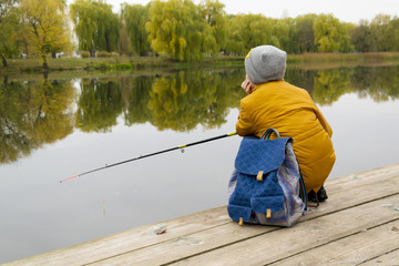 Fototapeta na wymiar Side view on boy in yellow jacket using fishing rod outdoors at pond during cool autumn day