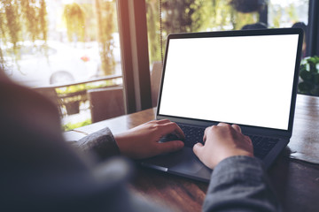 Mockup image of business woman using and typing on laptop with blank white screen and coffee cup on glass table in modern loft cafe