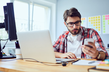 Focused young web developer checking his phone while working at his desk