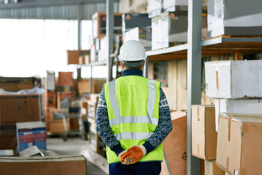 Rear view portrait of warehouse worker wearing hardhat and reflective jacket walking between storage shelves with boxes